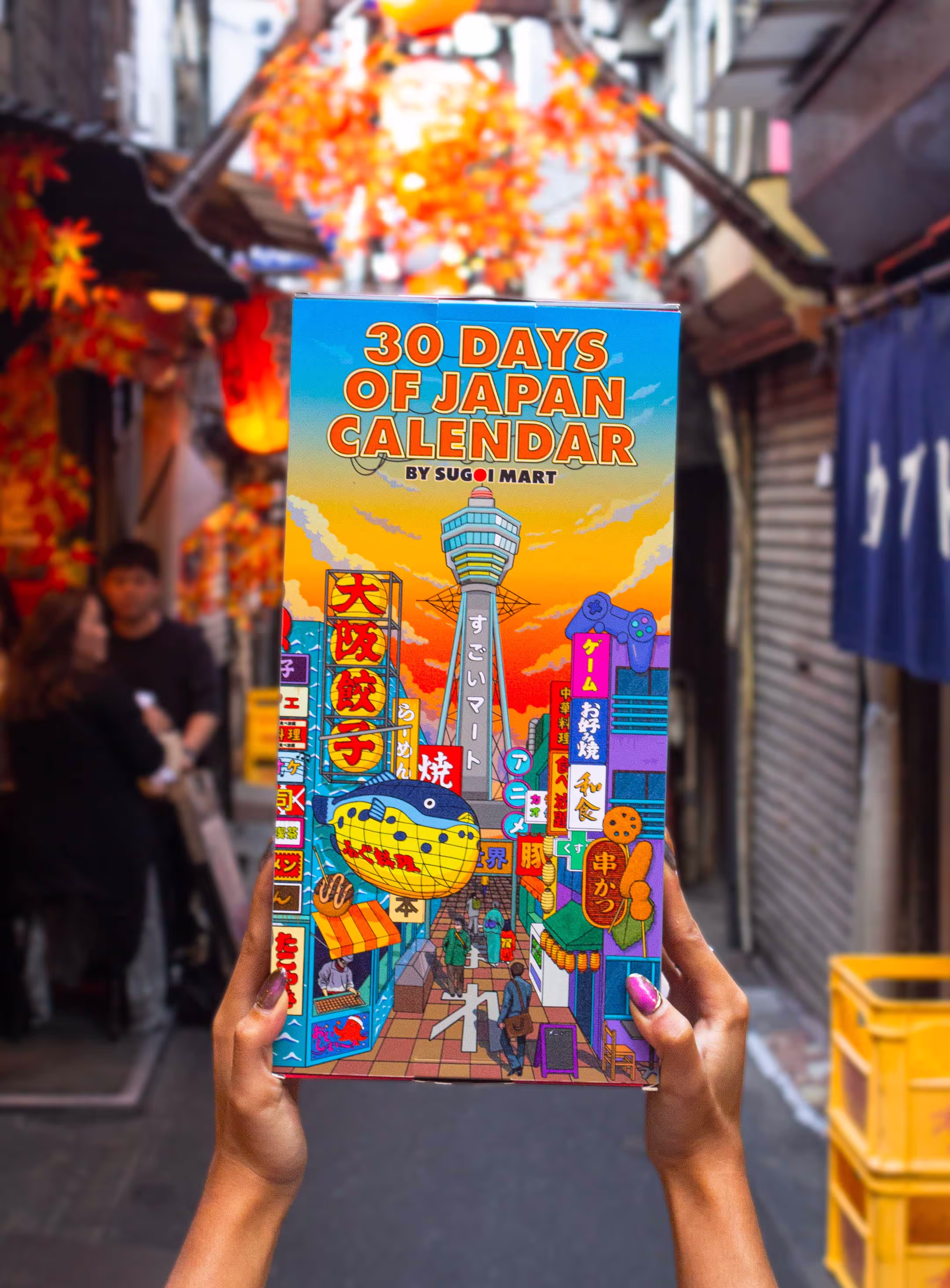 Hands holding up a vibrant and colorful package on a Japanese back alley. Showing the outside of the package. The package is an advent calendar but has a unique Japanese aesthetic to it, with all sorts of Japanese illustrations and drawings on it and has "30 days of Japan" written boldly on it.